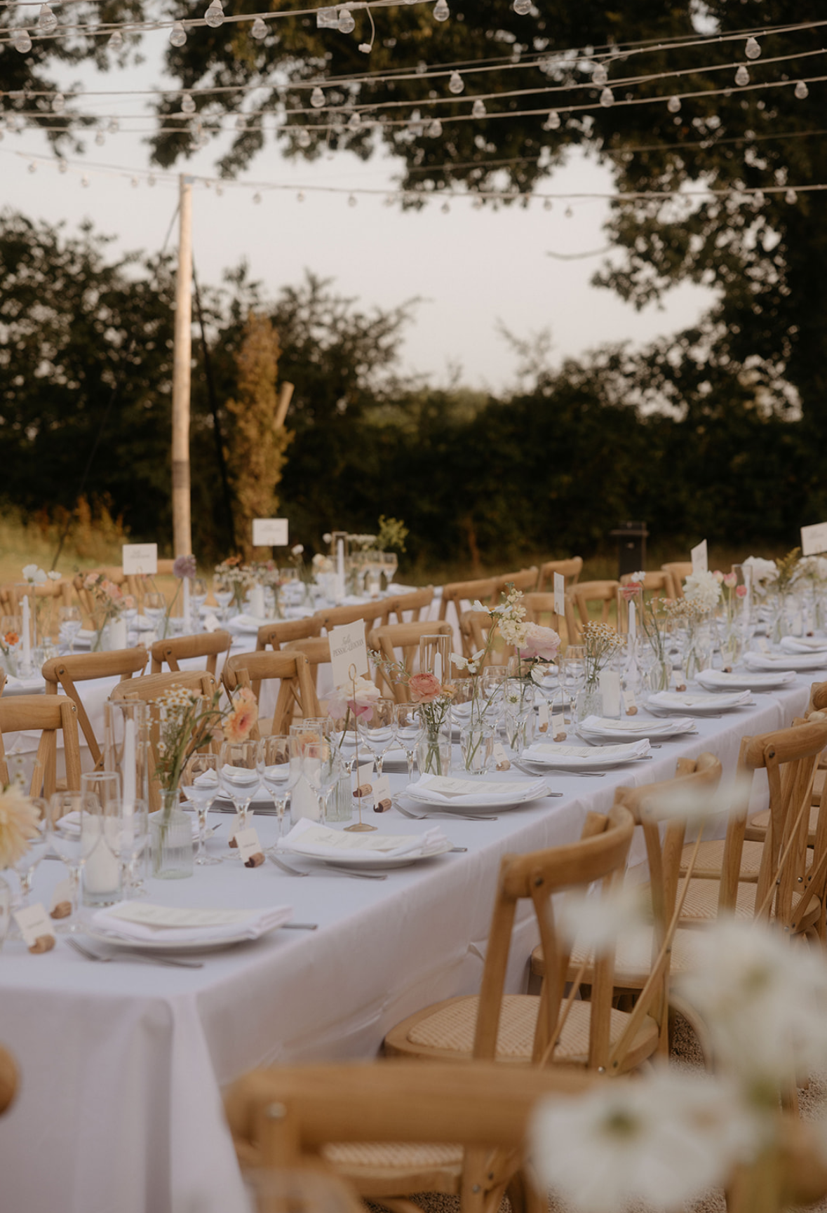 Longue table de mariage dressée en extérieur sous des guirlandes lumineuses
