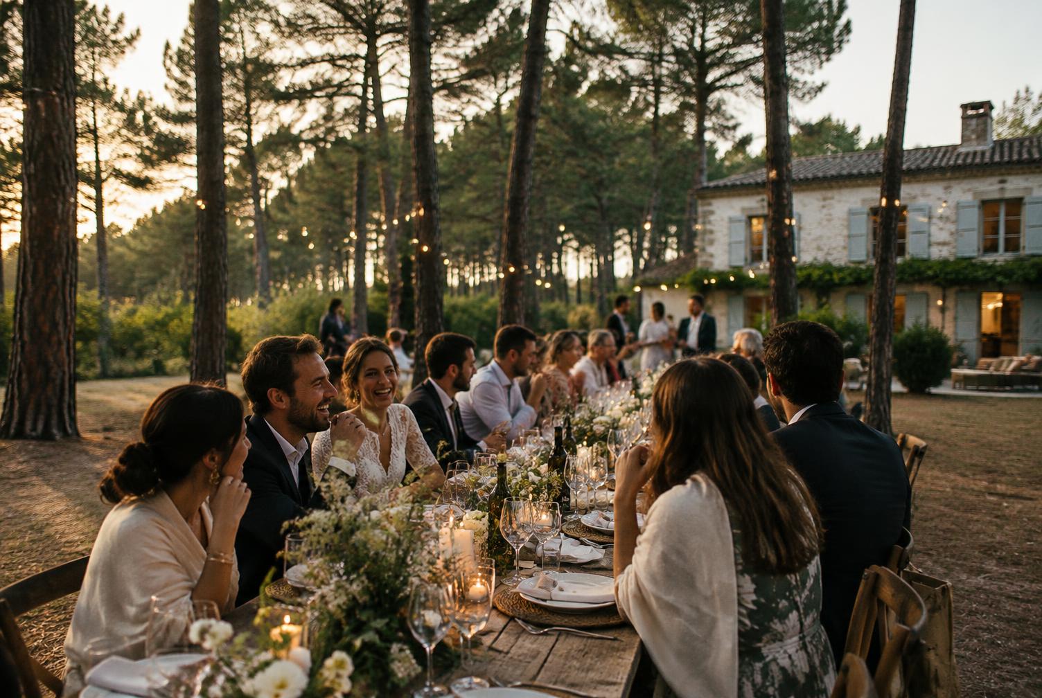 Mariés et invités qui rient autour d’une longue table en plein air